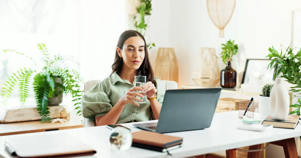 Water, glass and woman at laptop in home office for research, review and business plan for freelance project. Remote work, relax and consultant at desk with drink, computer and reading online article.