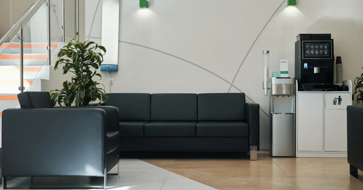 A modern office lobby features black seating, a water cooler station, and a tall indoor plant near a staircase.