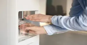 A person presses a button to fill a clear glass with water from a modern bottleless office water dispenser.