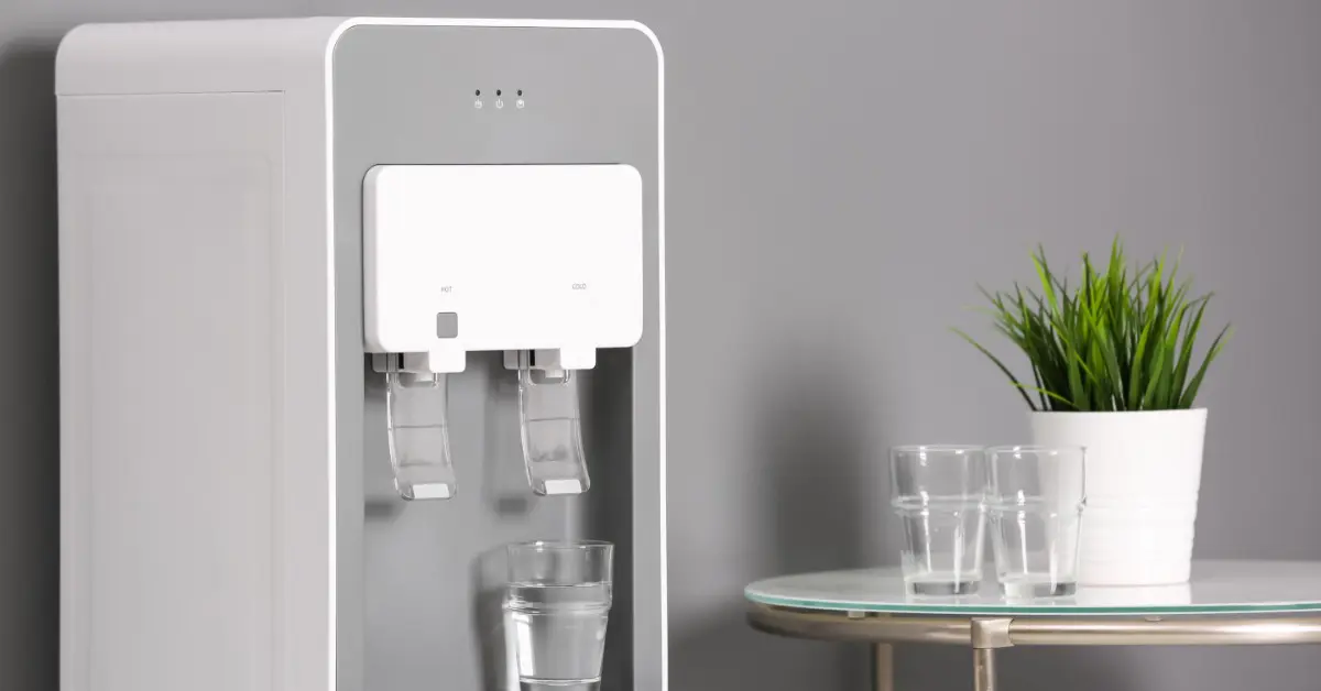 A modern bottleless water cooler stands beside a table with clear glasses and a small potted plant.