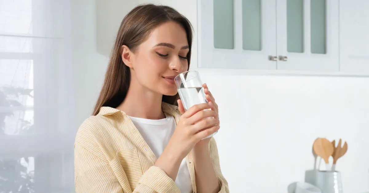 A young woman with brown hair in a yellow-striped shirt, drinking a glass of water while in a white kitchen.