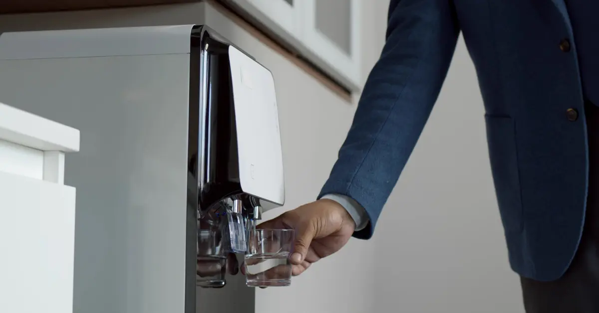 A close-up of a person in a suit filling a glass with water from a sleek bottleless office water cooler.
