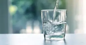 Clear water pours into a glass on a countertop, creating bubbles as it fills against a softly blurred background.