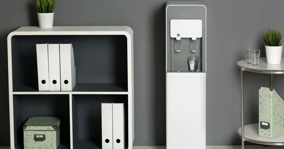 A bottleless water dispenser in an office. There are two men behind it, drinking water and talking with each other.