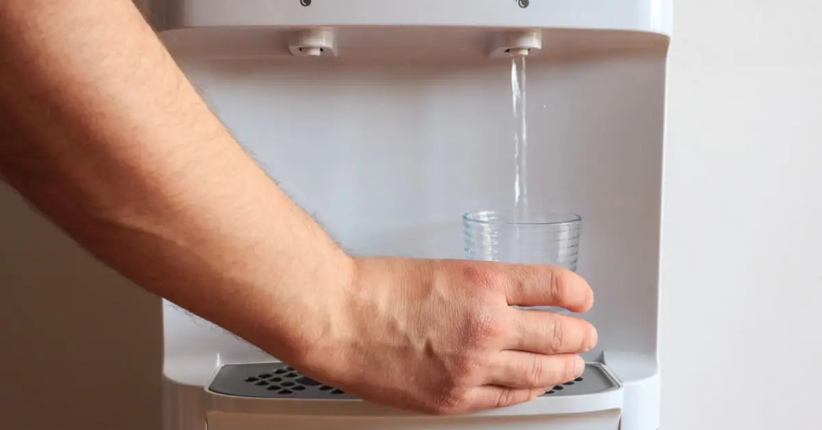 A bottleless water dispenser set up next to a small bookshelf and table. There are a few plants and file holders on them.