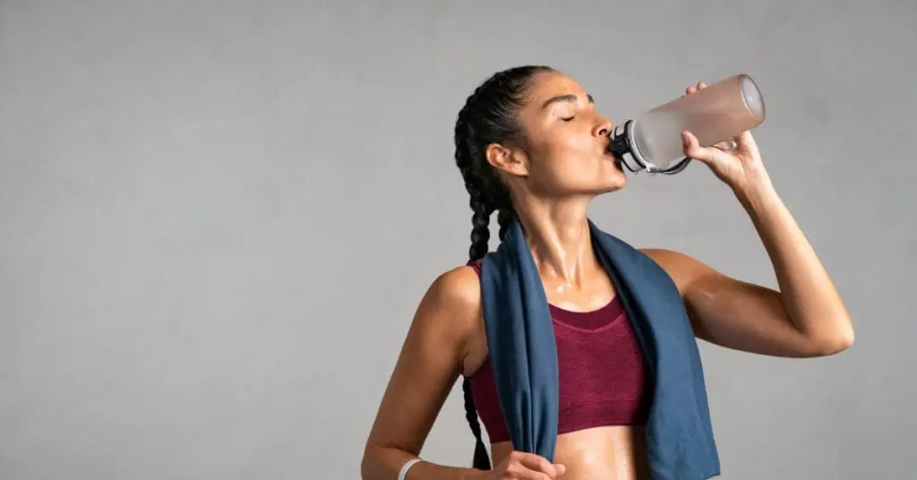 A woman in athletic gear drinking from a water bottle while holding a towel that's hung around her neck.