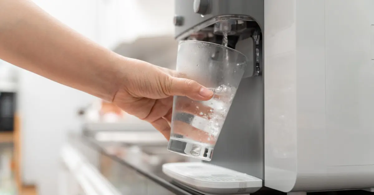 A hand holding a glass under a water cooler as clear drinking water pours from the dispenser into the cup.