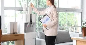 A businesswoman taking a paper cup from a water cooler in an office while holding a clipboard at her side.