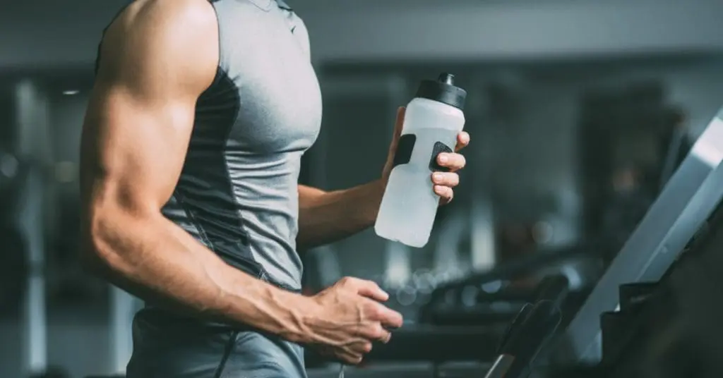 A man running on a treadmill at a gym while holding a water bottle during a steady indoor workout routine.
