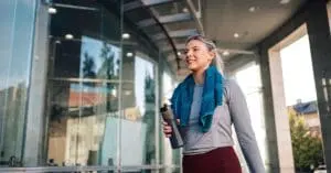 A woman in sports clothes leaving a gym holding a water bottle. She also has a towel around her neck.