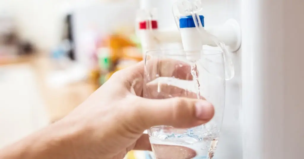 A person filling a glass with water from a countertop water cooler. They are using the cold dispenser to fill their glass.
