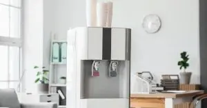 A modern water cooler standing in the middle of a bright office with a bunch of plastic cups stacked on top of it.