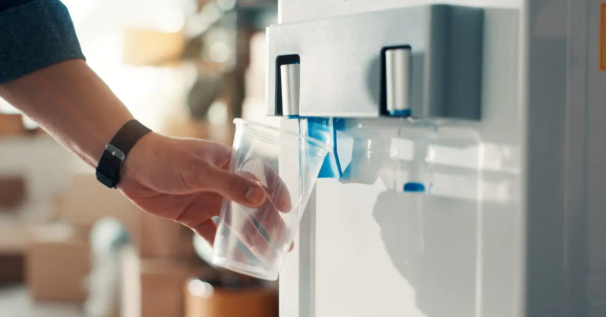 A hand holding a plastic cup under an office water dispenser while clear water is pouring from the spout.