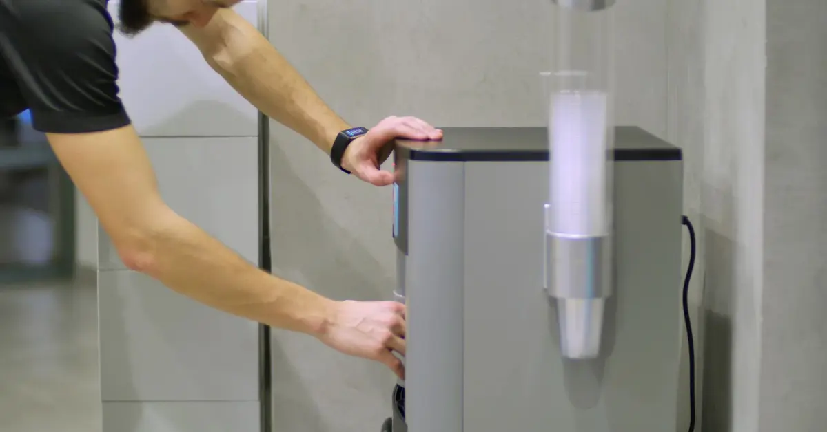 A man leaning over a gray water cooler, pressing the dispenser while one hand rests on the black top.