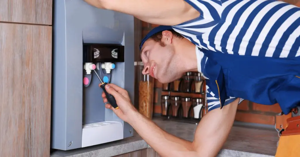 A technician repairing a water cooler in a kitchen with a screwdriver. He is hunched over, looking up at the dispenser.