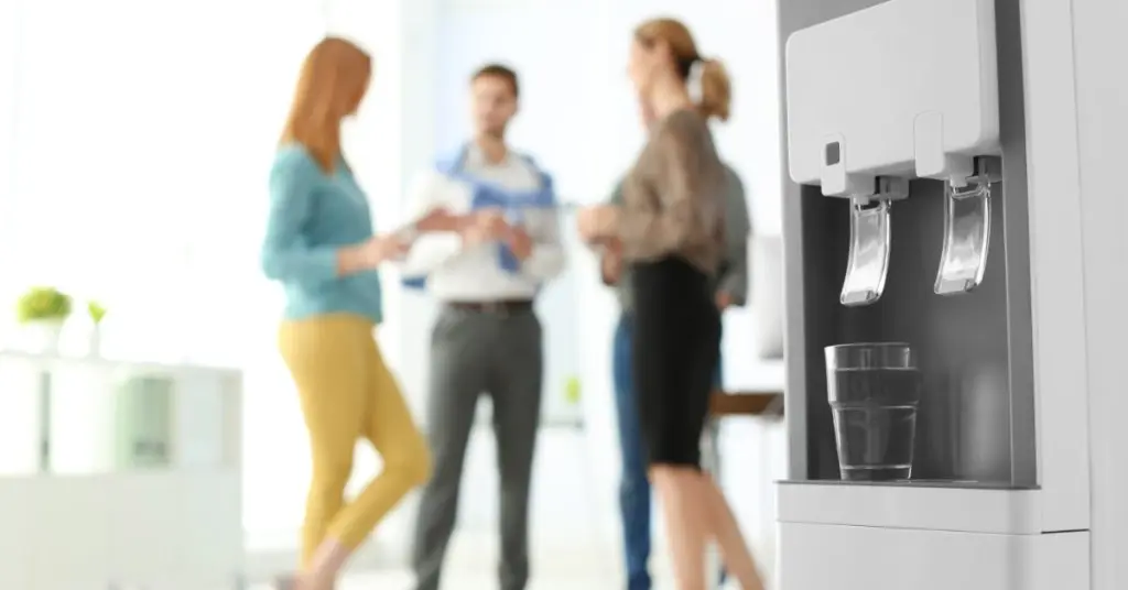 A close-up of a modern water cooler in an office space. There are people gathered together in the background.