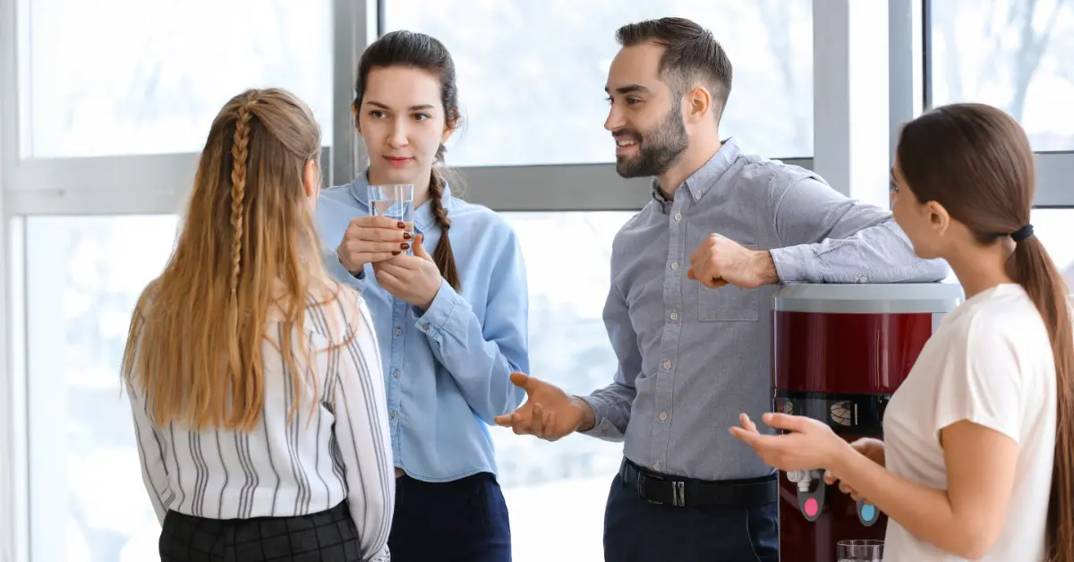 Four office workers standing together around a water cooler. One is holding a glass and another is leaning on the cooler.