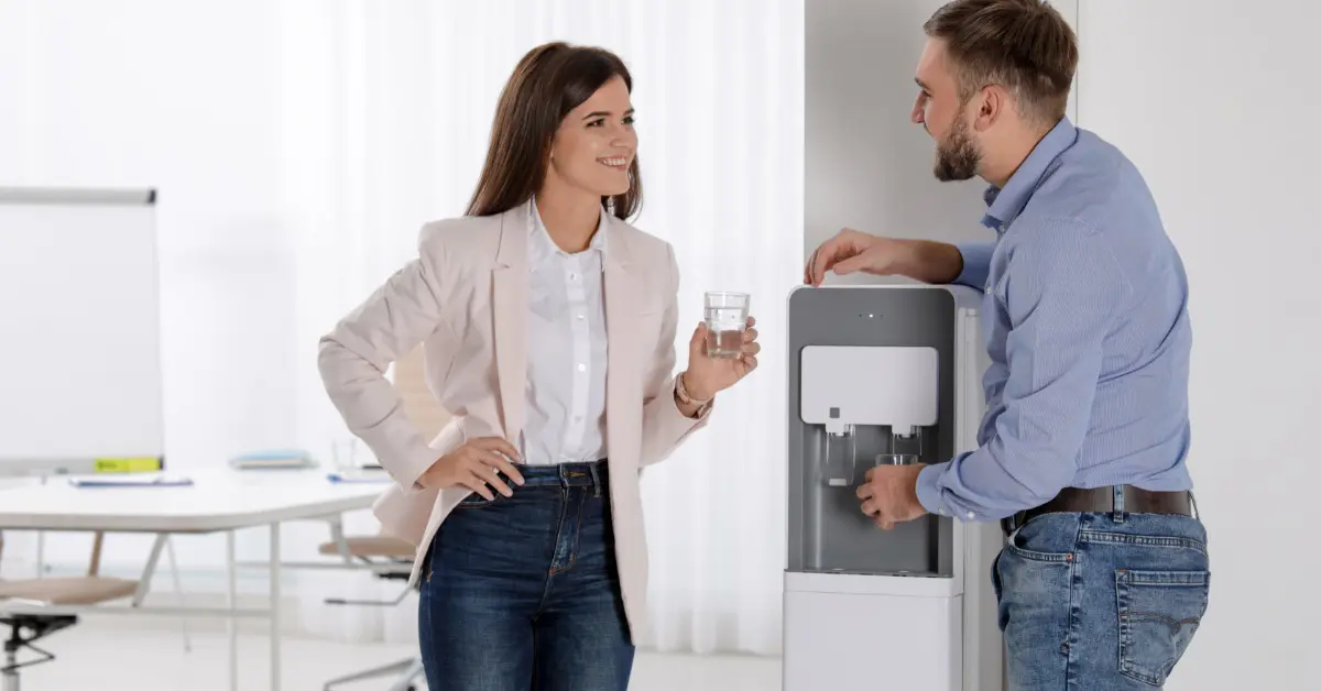 A close-up of two office workers standing together next to a water cooler. One is holding a cup and the other is getting water.