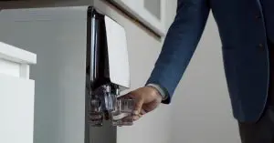 A person in a blue jacket holding a clear glass beneath a water dispenser while filling it with drinking water.