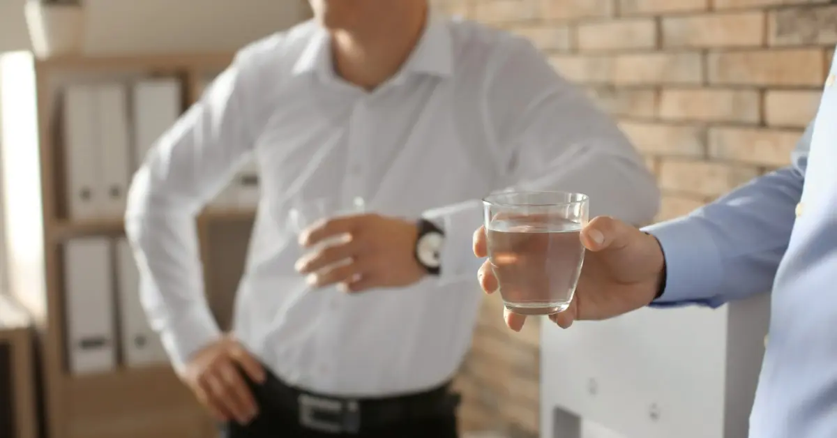 Two people standing near a water dispenser, each holding a clear glass of water in a bright office setting.