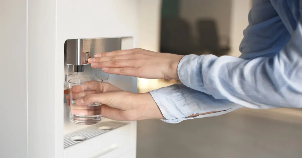 A person in a blue shirt holding a clear glass under a white water dispenser while pressing the dispensing button.