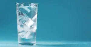 A clear glass full of ice cubes and water sits on a rough blue surface against a bright blue background.