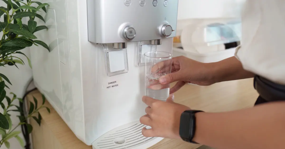 Someone filling a clear glass from a modern water dispenser in a bright kitchen, with a houseplant nearby.