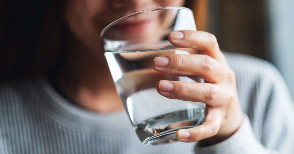  A woman bringing a clear glass full of water up to her lips. There is a bright light coming from behind her.