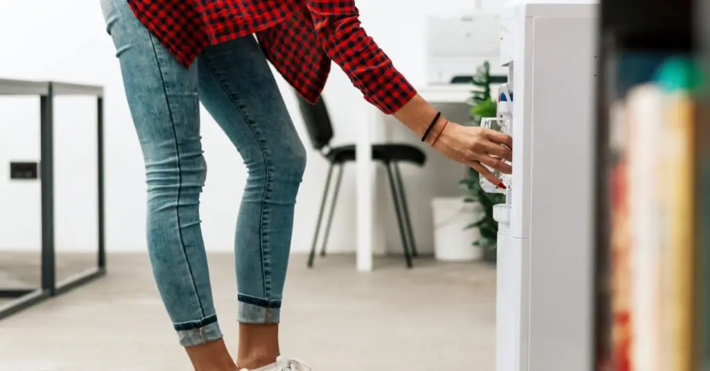A person in jeans and white sneakers filling a clear glass from a freestanding water dispenser in an office.