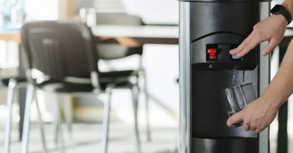 A person pressing the blue button on a black water cooler while holding a clear glass beneath the spout.