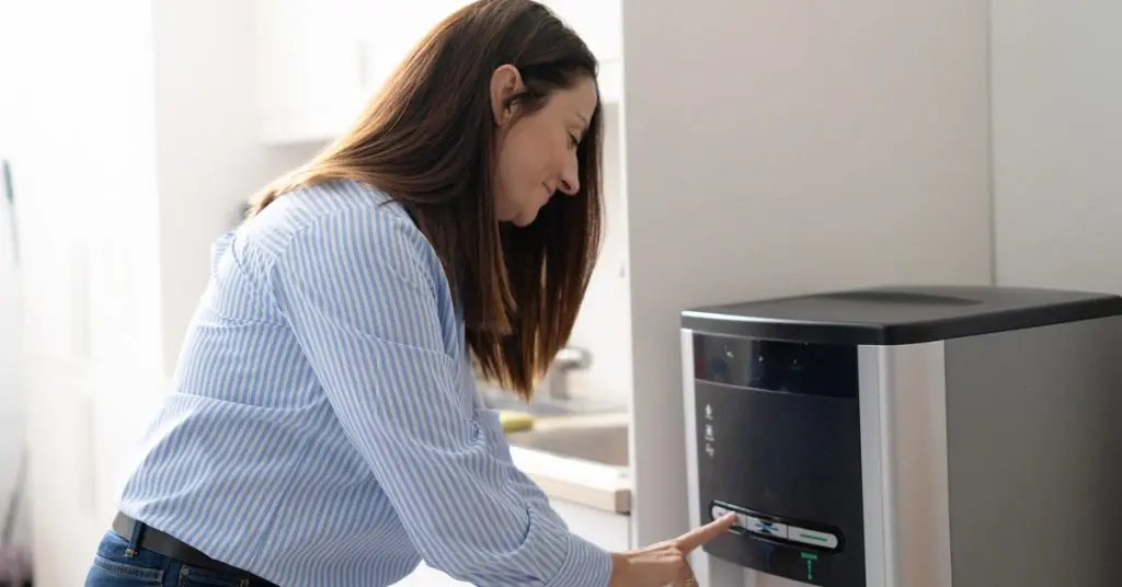 A woman pressing a button on a bottleless water dispenser while holding a small white cup beneath the spout.