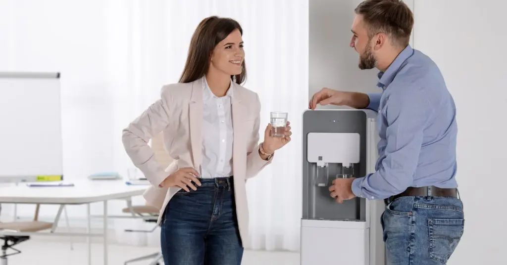 Two coworkers talking beside an office water dispenser while one holds a glass and the other fills a cup.