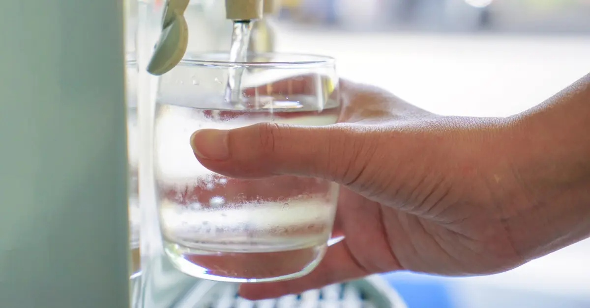 A hand holding a clear glass as water pours from a dispenser spout, filling the glass nearly to the top.