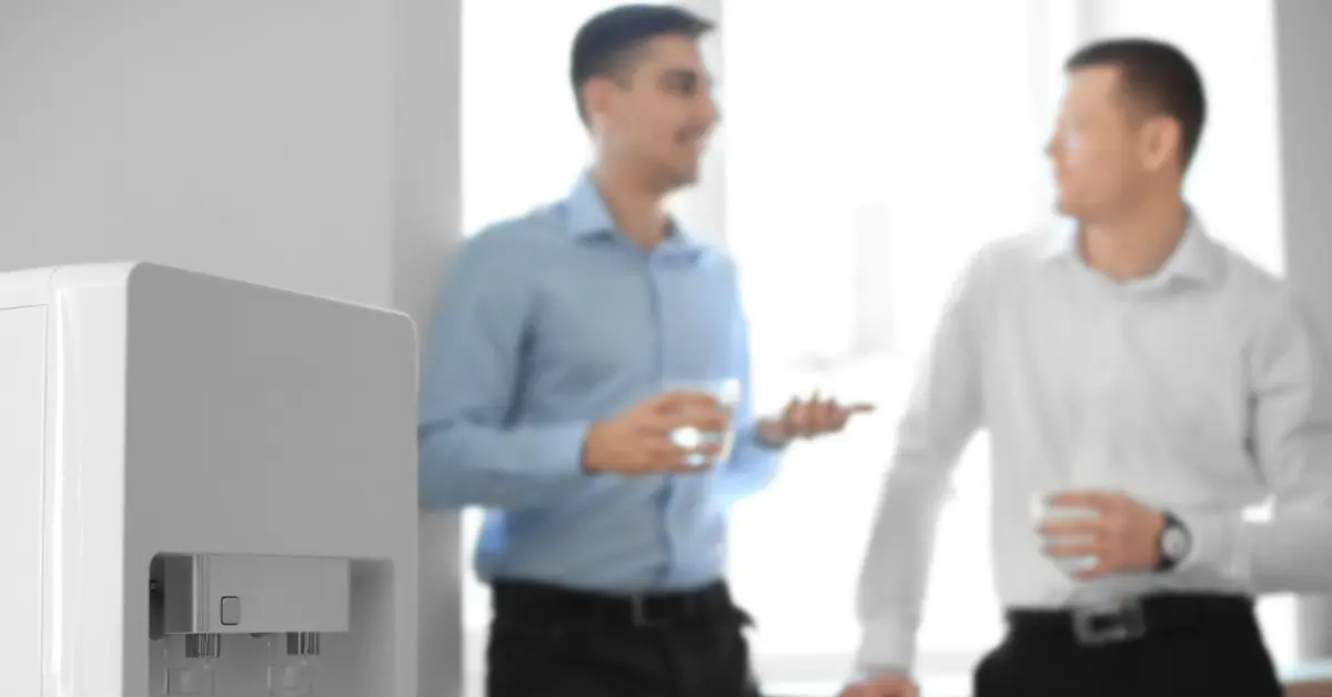 A white water dispenser in the foreground, with two coworkers holding cups while talking together in a bright office.