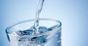Clear water being poured into a transparent drinking glass, creating swirling ripples and bubbles against a blue background.