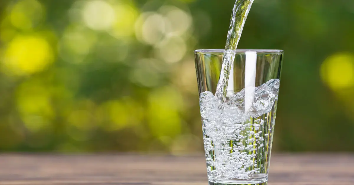 Clear water pouring into a drinking glass on a wooden table, creating bubbles and splashes against a blurred green background.